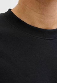 Črna bombažna majica s krožnim izrezom in gladko, mehko teksturo. Vidni so šivi vzdolž izreza.