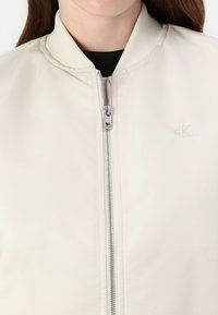 Kermakan värinen bomber-takki, jossa on ribbikraagi, etuvetoketju ja huomaamaton CK-logo rinnassa. Sileä, kevyt kangas.