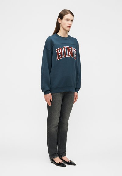 Ung person med rett brunt hår, iført en blå, oversized sweatshirt med teksten "BING", mørkegrå jeans og svarte spisse flate sko, stående.