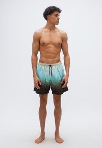 Άνδρας χωρίς πουκάμισο, όρθιος, ξυπόλητος, φοράει μαγιό με ombre χρώμα από πράσινο σε μαύρο και κορδόνι, κοιτώντας προς τα δεξιά του, μπροστά σε ένα απλό φόντο.