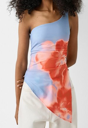 Femme portant un top tie-dye bleu et rouge à une épaule avec un ourlet asymétrique, associée à un pantalon blanc, debout devant un fond uni.