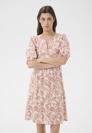 Jeune femme aux longs cheveux bruns portant une robe crème longueur genou avec un imprimé paisley rouge, debout bras croisés, expression neutre.