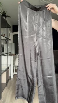 Szürke bársonyos, bővülő nadrág sima textúrával. Magas derék és elülső redők jellemzik. Az anyag finom fényű.