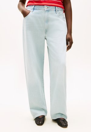 Inimene, kes kannab helesiniseid laia lõikega teksapükse, punaseid ja valgeid triibulist topi ning musti kootud lamedaid kingi, ühe käega taskus.