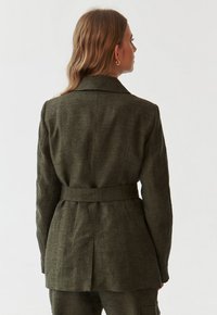 Grøn linnedjakke med en tætsiddende talje, oversized krave og sidesøm. Har lodret syning på ærmerne og en rygventilation.