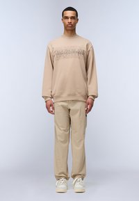 Felpa beige con stampa grafica, abbinata a pantaloni color beige chiaro e sneakers bianche. Presenta una vestibilità comoda e polsini a coste.