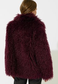 Giacca in pelliccia sintetica bordeaux con una vestibilità oversize, texture shaggy e colletto spesso, dotata di maniche lunghe e un aspetto morbido e peloso.