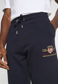 Námořnické tepláky s pružným pasem a zlatými doplňky. Na nich je vyšitý logo "GANT" a "AMERICAN SPORTSWEAR" v červené a zlaté barvě.