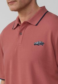 Man in een gedempte rode polo met donkerblauwe rand op de kraag en een klein geborduurd donkerblauw vintage auto op de borst.
