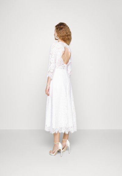 Hvid blondekjole med trekvartærmer, åben ryg og knælang skørt. Bæres med strappy hvide højhælede sandaler.