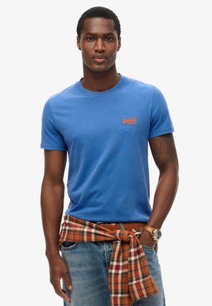 Blå bomulds T-shirt med rund halsudskæring og korte ærmer, med et orange logo. En plaidskjorte er bundet om taljen.