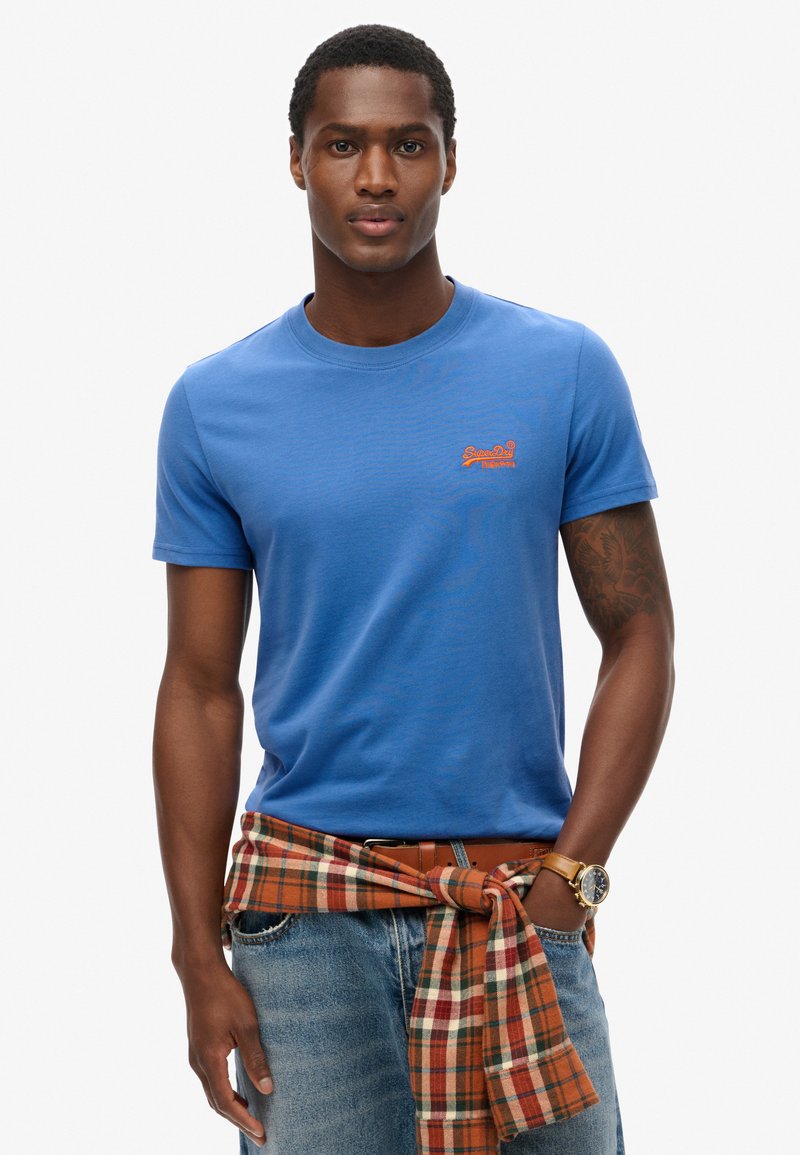Camiseta de algodón azul con cuello redondo y mangas cortas, que presenta un logo naranja. Una camisa de cuadros está anudada en la cintura.