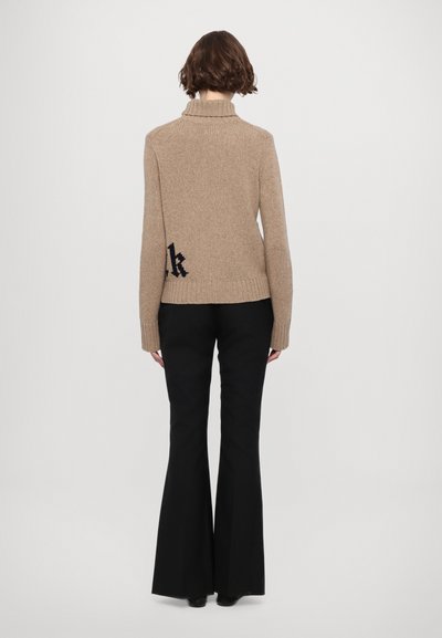 Beige gestrickter Rollkragenpullover mit geripptem Saum und Bündchen, mit einem schwarzen gestickten Detail auf dem unteren Rücken. Kombiniert mit schwarzen Schlaghosen.