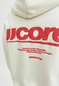 Sudadera con capucha crema con texto grande rojo "JJcore" y texto rojo más pequeño debajo que dice "Innovación Creativa Desde 1990" y el logo de "jack&jones".