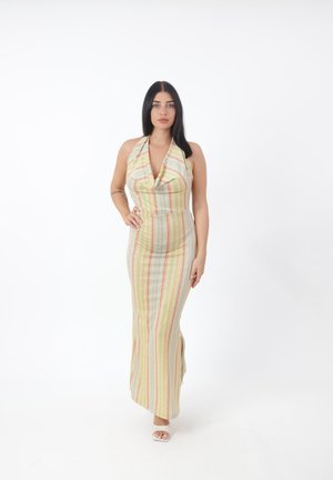 Kobieta o długich ciemnych włosach, ubrana w bezrękawnikową, paseczkową sukienkę maxi z dekoltem halter, stojąca z jedną ręką na biodrze na białym tle.