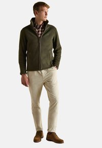 Olivovo zelená fleecová bunda s vysokým golierom, zipsom a bočnými vreckami, skombinovaná s krémovými kordovými nohavicami a hnedými šnurovacími čižmami.