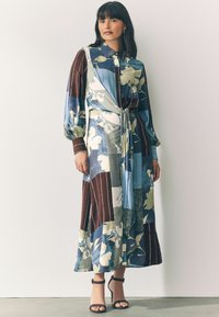 Blumenmusterkleid mit langen Ärmeln, gemischte Stoffpaneele in Blau, Grün und Braun. Mit einem gebundenen Taillendetail, kombiniert mit schwarzen, hohen Sandalen.