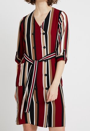 Vrouw draagt een knielange jurk met verticale strepen in rood, zwart, wit en bruin, met knopen en een bijpassende gestreepte riem.