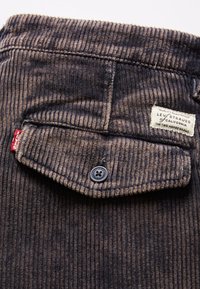 Tissu en velours côtelé brun foncé avec une poche à rabat boutonnée, une étiquette rouge Levi's et une étiquette blanche de marque cousues au-dessus de la poche.