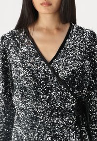 Must musträtiga ülemine osa, mis on kaetud läikivate hõbedaste sädelevate sequinitega, sisaldab V-dekolteed ja musta nööri sulgemist. Sile tekstuur erinevate valguse peegeldustega.