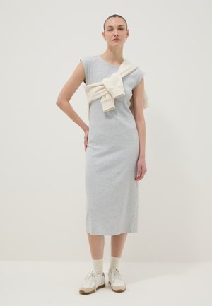 Vrouw staand, gekleed in een mouwloze grijze midi-jurk met een witte trui om haar schouders gestrikt en witte sneakers.