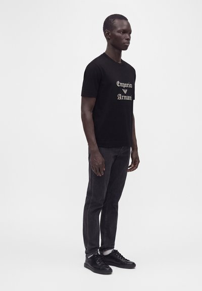 Άνδρας που στέκεται, φοράει μαύρο T-shirt Emporio Armani, σκούρο τζιν, λευκές κάλτσες και μαύρα αθλητικά παπούτσια, σε απλό λευκό φόντο.