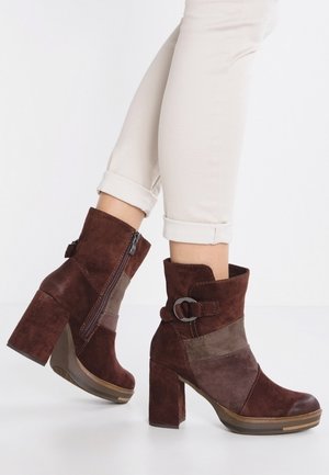 Bottines en daim marron avec talons blocs, fermeture éclair latérale et boucle décorative, portées avec un pantalon beige clair retroussé.