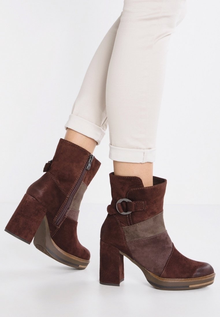 Bottines en daim marron avec talons blocs, fermeture éclair latérale et boucle décorative, portées avec un pantalon beige clair retroussé.
