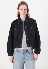 Giubbotto in denim nero con taglio corto, due tasche sul petto e chiusura a bottoni. Abbinato a jeans a vita alta di colore blu chiaro.