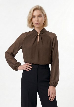 Blonde Frau trägt eine braune, transparente Bluse mit langen Ärmeln, einem Ausschnitt mit Schlitz und goldener Spange, kombiniert mit hoch taillierter schwarzer Hose.