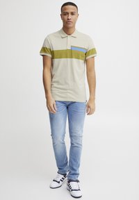 Beige polo skjorte med grønne og blå horisontale striper. Bomullsmateriale, avslappet passform, kombinert med lyseblå jeans og hvite joggesko.