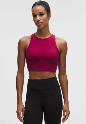 Frau mit dunklen Haaren im hohen Dutt, trägt ein ärmelloses dunkelrosa Crop-Top und hochtaillierte schwarze Leggings vor einem einfachen hellen Hintergrund.