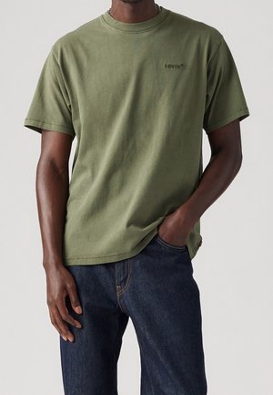 Man in een olijfgroene Levi's t-shirt en donkerblauwe spijkerbroek, met één hand in zijn zak, staand tegen een effen achtergrond.