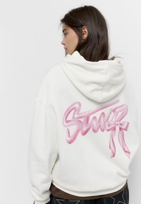 Hvid hættetrøje med et pink spraymalet "Stuer" logo og sløjfedetalje på ryggen. Har en snoretrukket hætte og løs pasform.