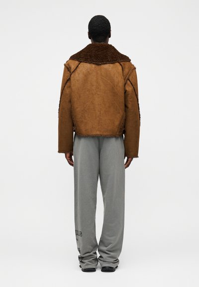 Καφέ δερμάτινο μπουφάν με καφέ γούνινο γιακά, σε τετράγωνο σχήμα με εμφανείς ραφές, σε συνδυασμό με ανοιχτό γκρι, φαρδιά παντελόνια.
