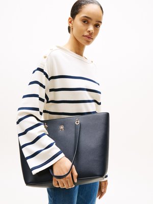 Bolso tote de cuero navy con un acabado texturizado, que presenta herrajes dorados y un logo. El modelo lleva una camisa a rayas con un escote barco.