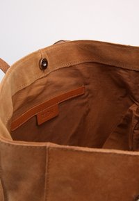 Binnenkant van een bruine suede totebag met een drukknoopsluiting en een binnenzak, voorzien van een leren label met de opdruk "ZIGN".