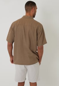 Kortærmet brun skjorte i struktureret stof. Har en afslappet pasform, krave og detalje ved ryggen. Bæres med lyse shorts.