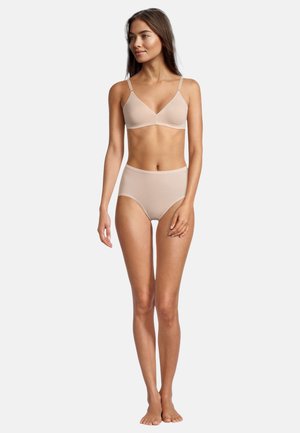 Frau steht in beige nahtlosem BH und hoch taillierter Unterwäsche und blickt nach links vor einem schlichten weißen Hintergrund.