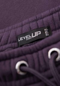 Fialový textilní oděv s texturovaným páskem, stříbrnými průchodkami a černou nálepkou se slovy "LEVEL UP" a "ombre" v bílém písmu.