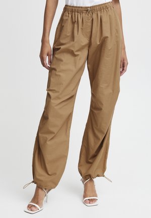 Brown, lightweight, relaxed-fit trousers with an elastic waistband, adjustable drawstring, and cinched hems, paired with white strappy heels.