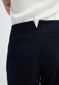 Close-up van marineblauwe pantalon met een V-vormige inkeping op de tailleband, gedragen met een witte gebreide top met structuur die erin gestoken is.