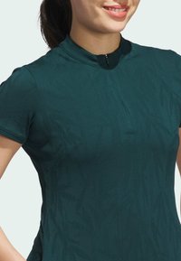 Mörk teal kortärmad topp med hög krage, som har ett subtilt geometriskt mönster och en partiell framdragkedja. Slät, figurnära textur.