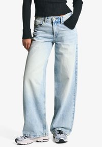 Svijetloplave široke traperice od denima, s izbljeđenim pranjem, visokim strukom i vidljivim šavovima. Uparene sa srebrnim sportskim tenisicama.