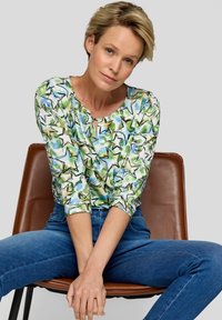 Bunt gemusterte Bluse mit einem blättrigen Sternenmuster, runder Ausschnitt und trois Viertelärmeln. Getragen mit blauen Jeans.