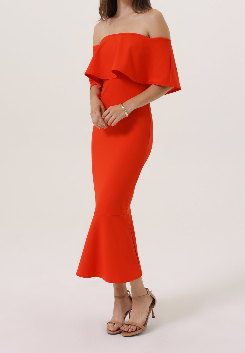 Femme portant une robe midi ajustée rouge vif à épaules dénudées, avec un ourlet évasé et des sandales à talons beiges, debout devant un fond uni.