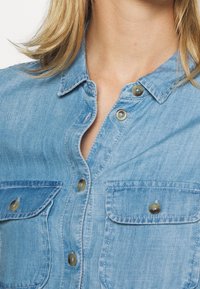 Vaaleansininen farkkupaita, jossa on napitettava etuosa, kaksi rintataskua ja pyöristetty kaulus. Kangas näyttää hieman teksturoidulta.