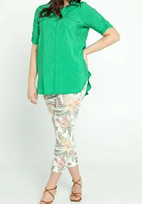 Donna che indossa una camicia verde a bottoni, pantaloni bianchi cropped con una stampa floreale e sandali marroni con delle cinghie, in piedi con una mano sulla vita.