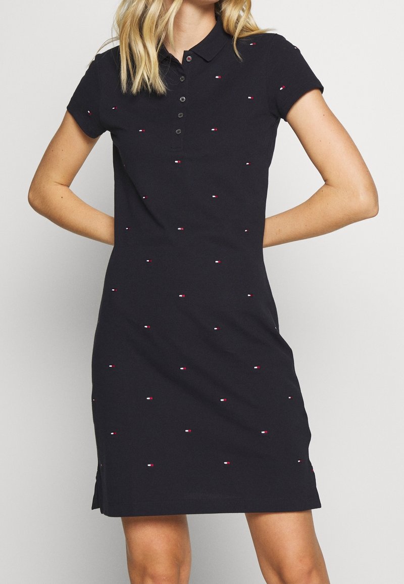 Femme portant une robe polo noire à manches courtes, col boutonné, et une petite broderie de logo en rouge, blanc et bleu répartie sur toute la surface.