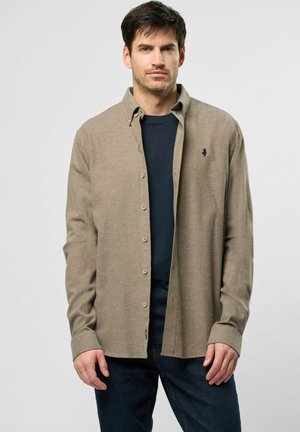 Beige knapp skjorte med lange ermer, med en subtil tekstur og lille logo på brystet, båret over en mørkeblå skjorte og jeans.